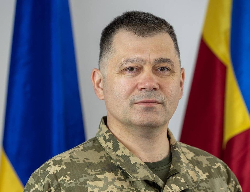 Brigaadikindral Poti portree õlgadeni. Tumedate lühikeste juustega laigulises riietuses mees seisab lippude ees.