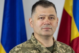 Brigaadikindral Poti portree õlgadeni. Tumedate lühikeste juustega laigulises riietuses mees seisab lippude ees.